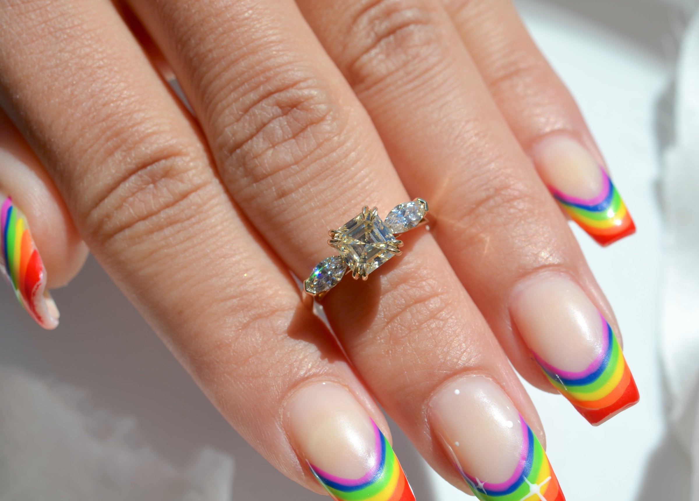 A custom diamond engagement ring by Emma Brooke, shown on a models hand. Designed and made in Portland, OR, this ring features a large antique Asscher square diamond. The model has a rainbow French-tipped manicure.