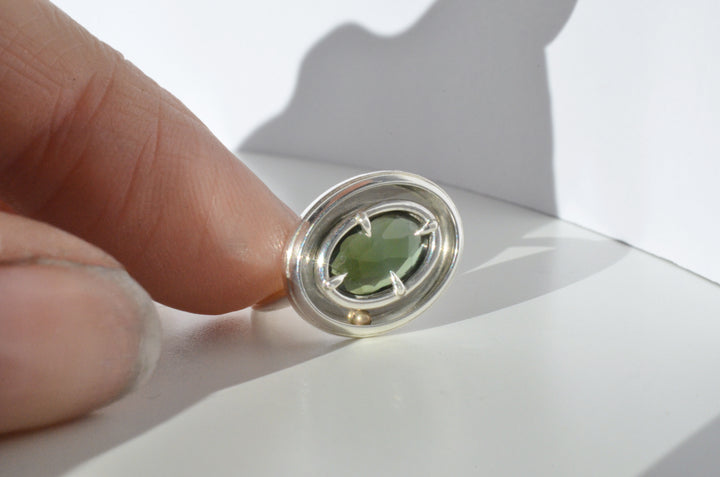A dynamic shot showing the kinetic green tourmaline orbit ring about to be set into motion. The rolling gold ball inside the ring is shifted to one side, by a hand tilting the ring to the side.