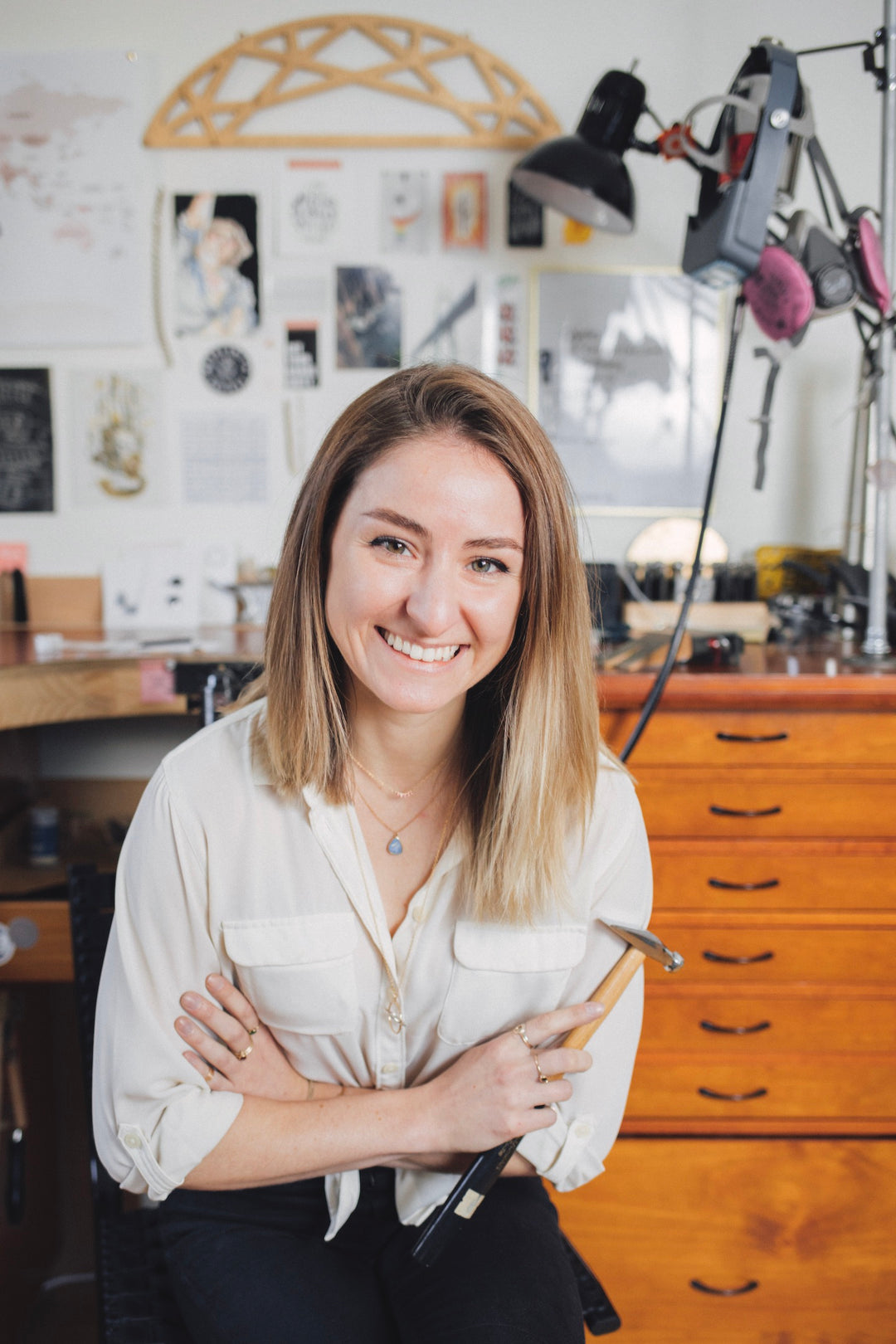 Smiling photo of portland custom jeweler Emma Brooke at custom jeweler workbench, wearing a white shirt and holding a hammer
