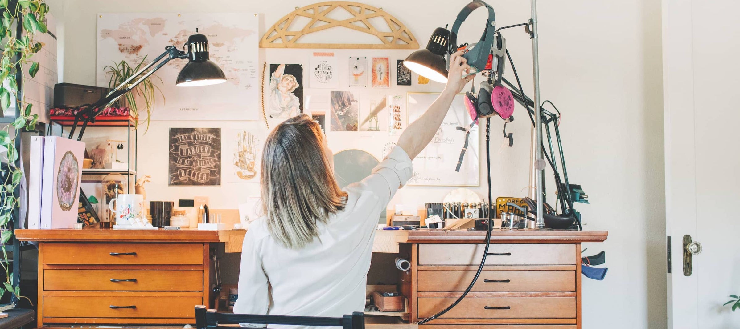 a photo of jewelry artist Emma Brooke from behind, sitting at her custom jewelers workbench. The bench is surrounded by plants, jewelry making tools, and  a collage of wall art. 