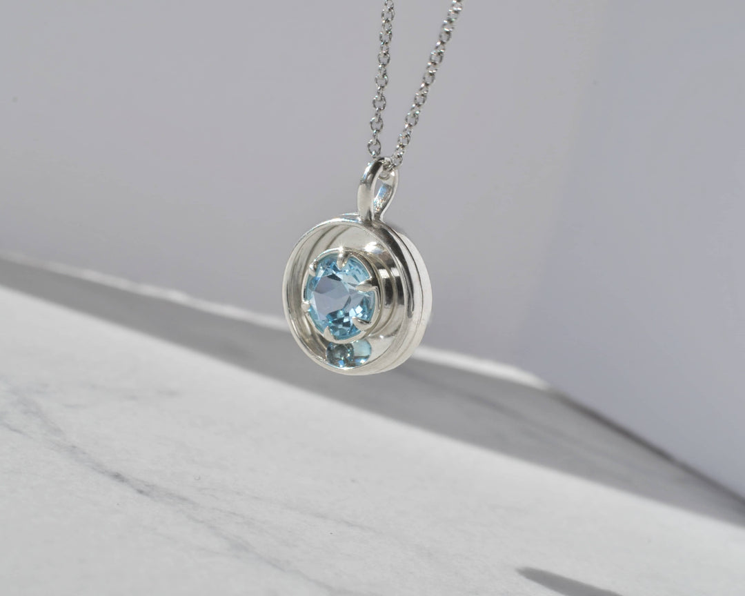Silver necklace with a pendant featuring blue gemstones suspended in sunlight against a white marble background