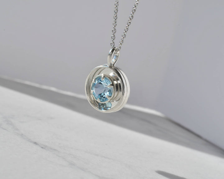 Silver necklace with a pendant featuring blue gemstones suspended in sunlight against a white marble background