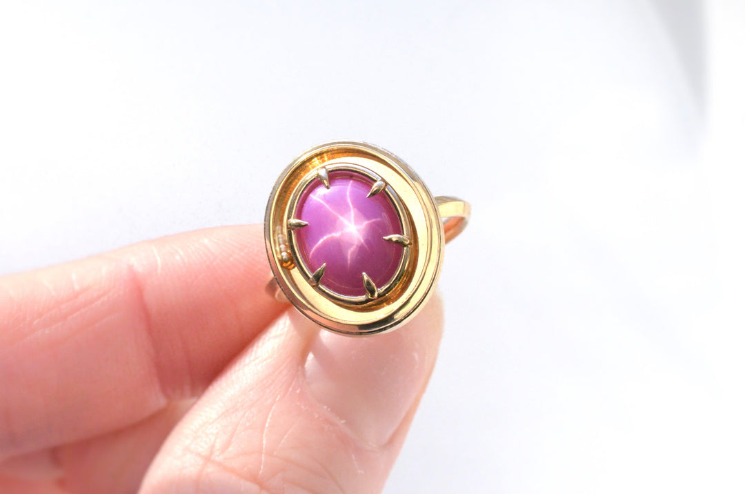 A star ruby solitaire orbit ring by fine jeweler Emma Brooke, featuring a kinetic rolling ball for fidgeting or meditation. The photo shows the ring in sunlight with a white background, and shows the ball in motion. The ring is yellow gold, with a hot pink ruby.