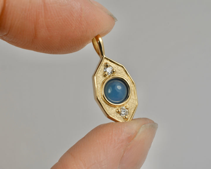 A gold pendant with a rolling blue spherical stone and two small white diamonds, held between fingers against a light gray background.
