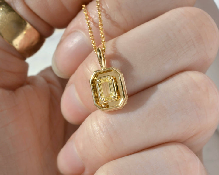 A close-up of a hand holding a gold pendant necklace with an emerald-cut yellow gemstone.