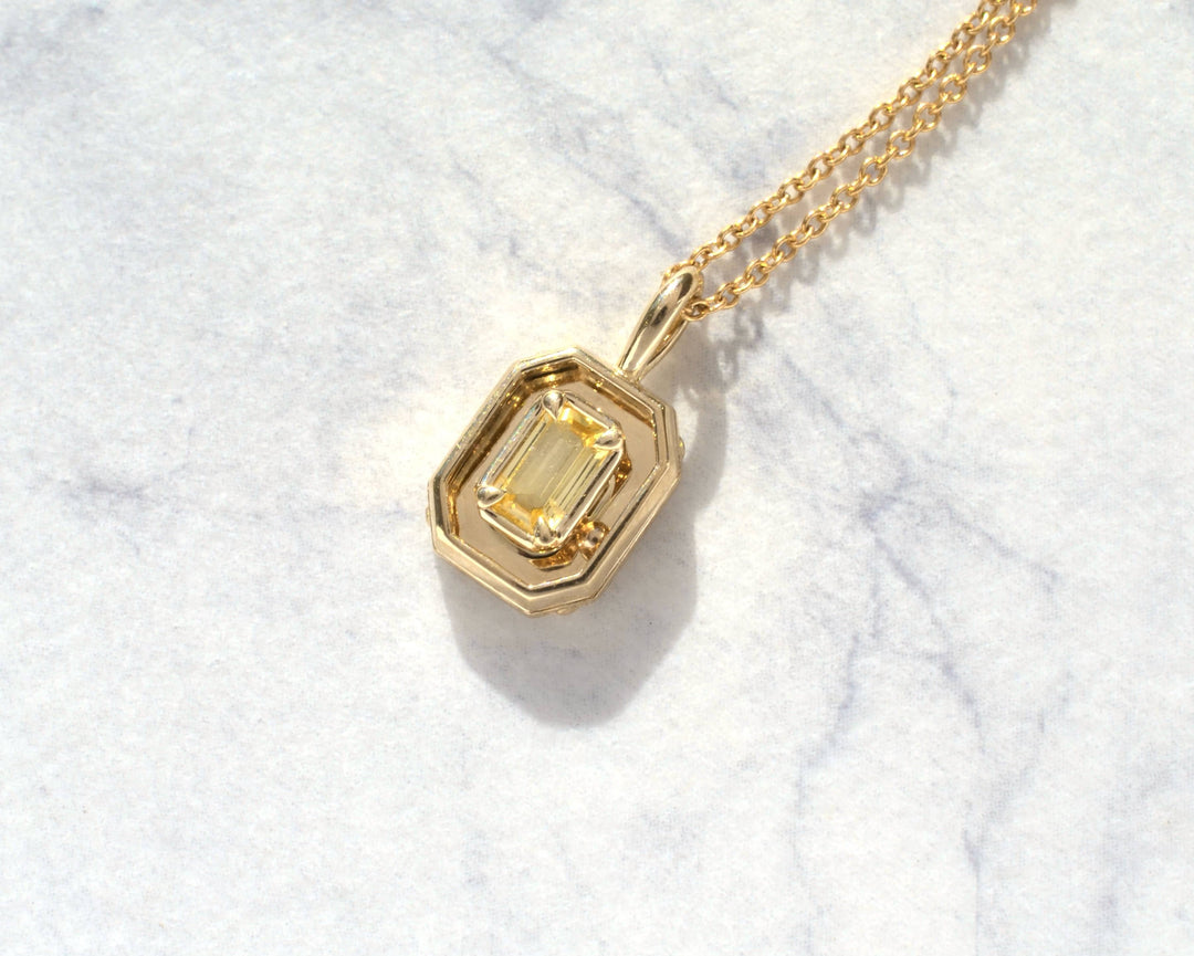 A gold pendant necklace featuring a yellow emerald-cut gemstone in a geometric bezel setting, resting on a white marble surface.