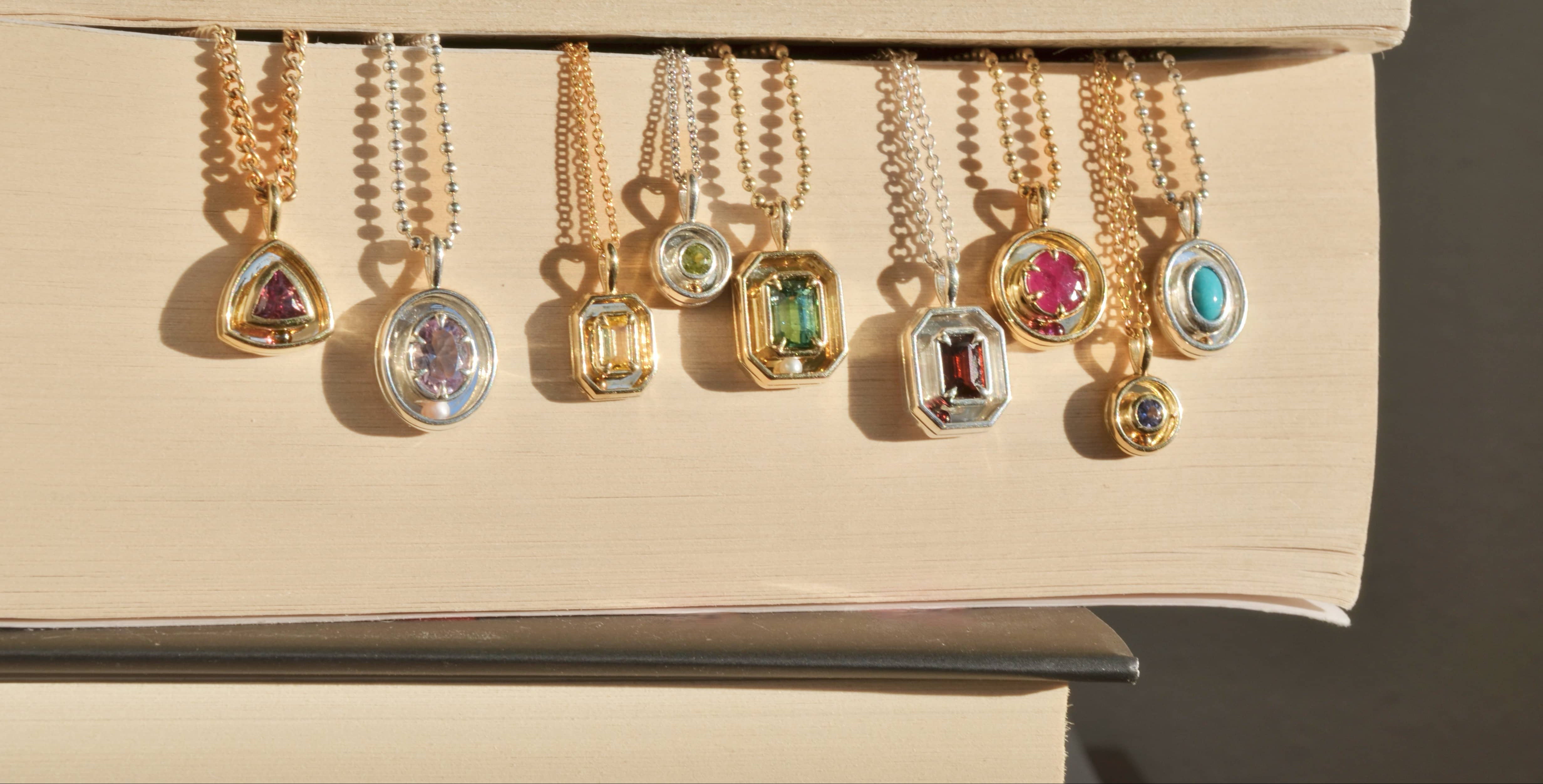 9 kinetic "orbit style" necklaces with colorful gemstone pendants are displayed hanging from the pages of stacked books.