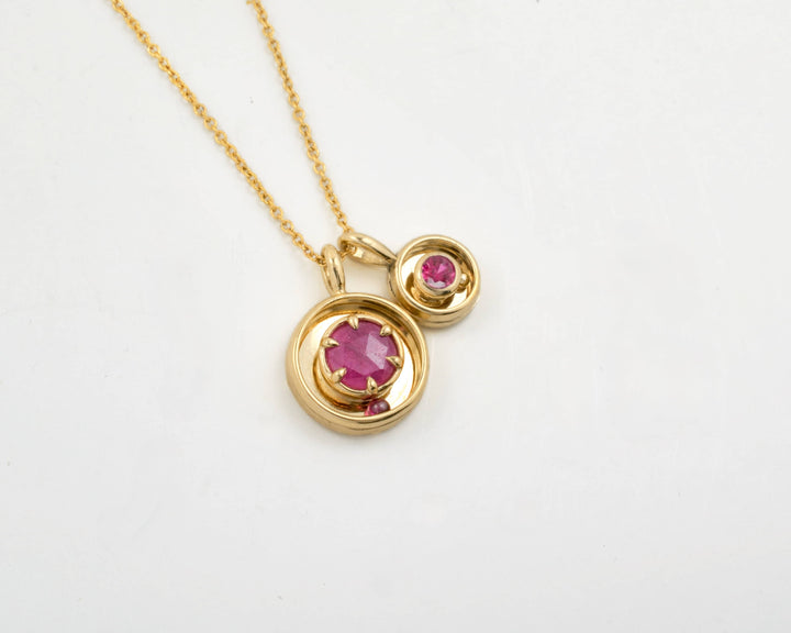 The Orbit Pendant, and the smaller Orbit birthstone charm side by side, both in yellow gold and ruby