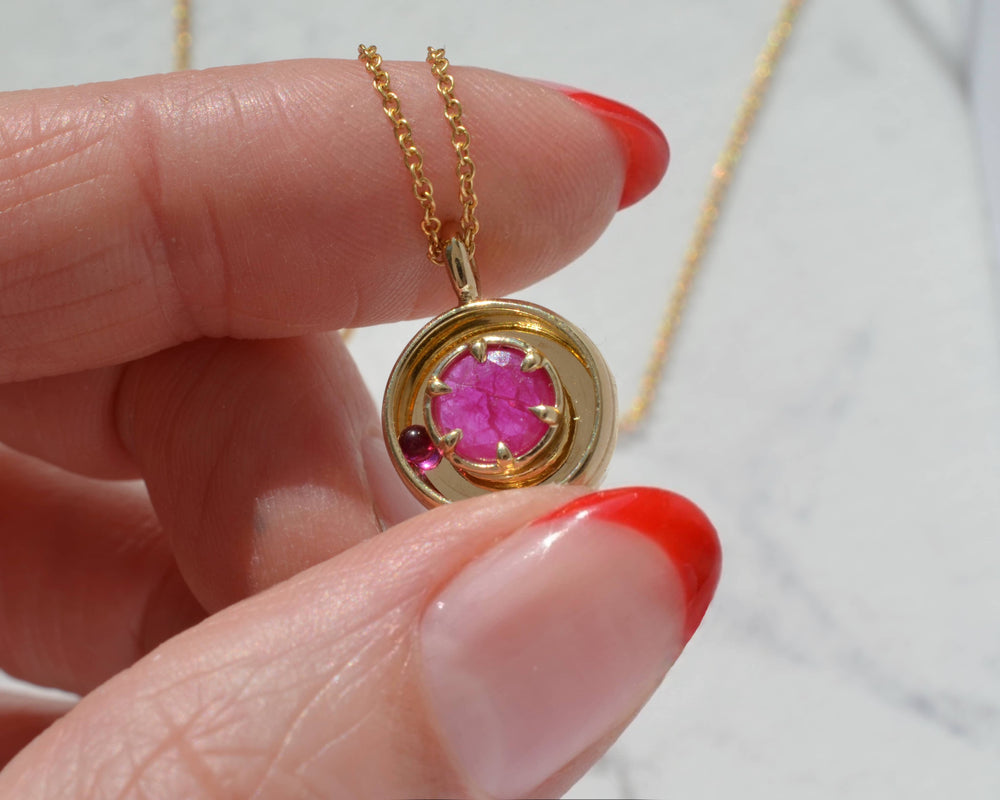 Gold pendant with a hot pink ruby and moving ball, held by a hand with a red tipped manicure