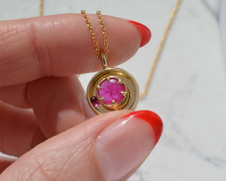 Gold pendant with a hot pink ruby and moving ball, held by a hand with a red tipped manicure