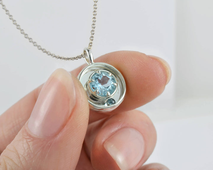 Silver kinetic rolling ball necklace with a blue gemstone held by a hand against a white background