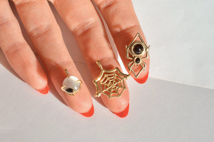 3 solid gold halloween themed charms, each resting on one fingertip of a red-manicured hand. the charms include a googly eye, a spider web, and a spider, and are all kinetic.