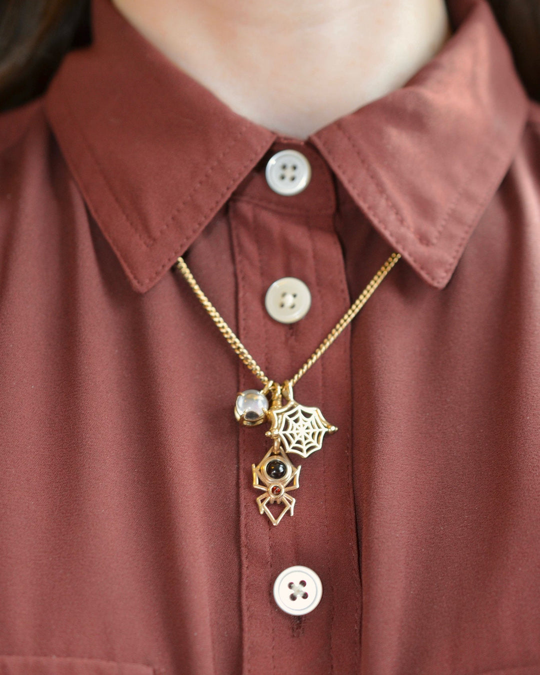 3 14k gold halloween themed charms including an eyeball, a spider web, and a spider, modeled against a burgundy red button-down shirt.