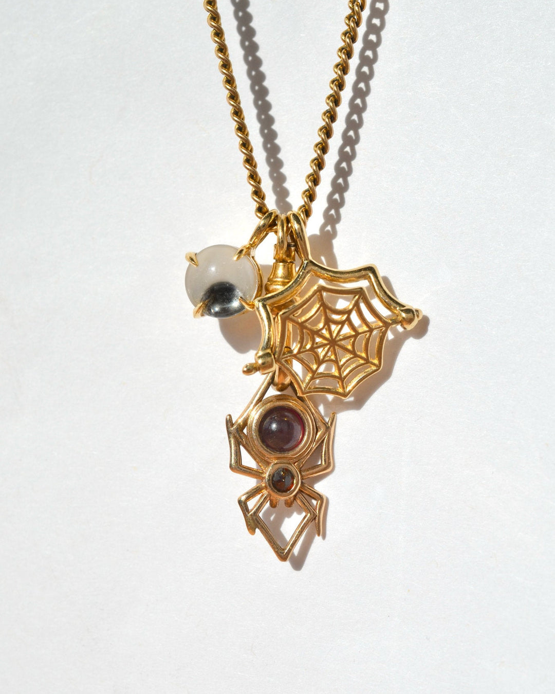 A gold-toned charm necklace featuring a spider, a spider web, and a googly eye charm, shown on a plain white background. the charms are all kinetic and can move.