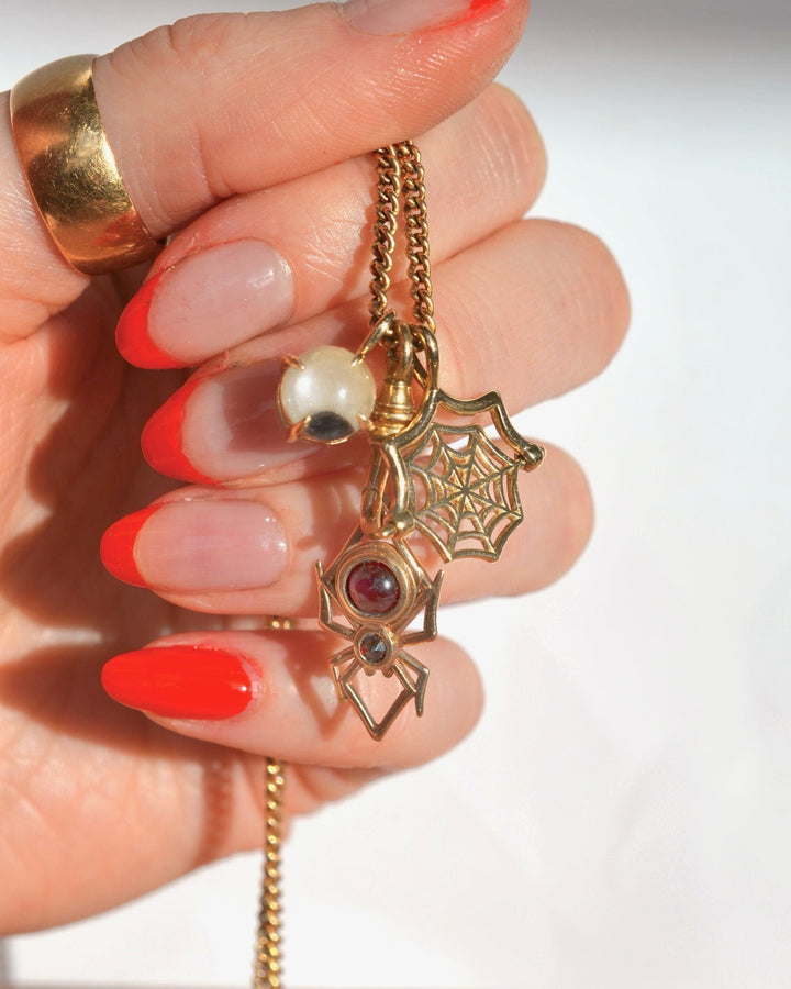 A close-up of a hand with red-painted nails holding a gold-tone chain necklace with halloween-themed charms, including a spider web pendant, a spider silhouette charm, and an eyeball charm.