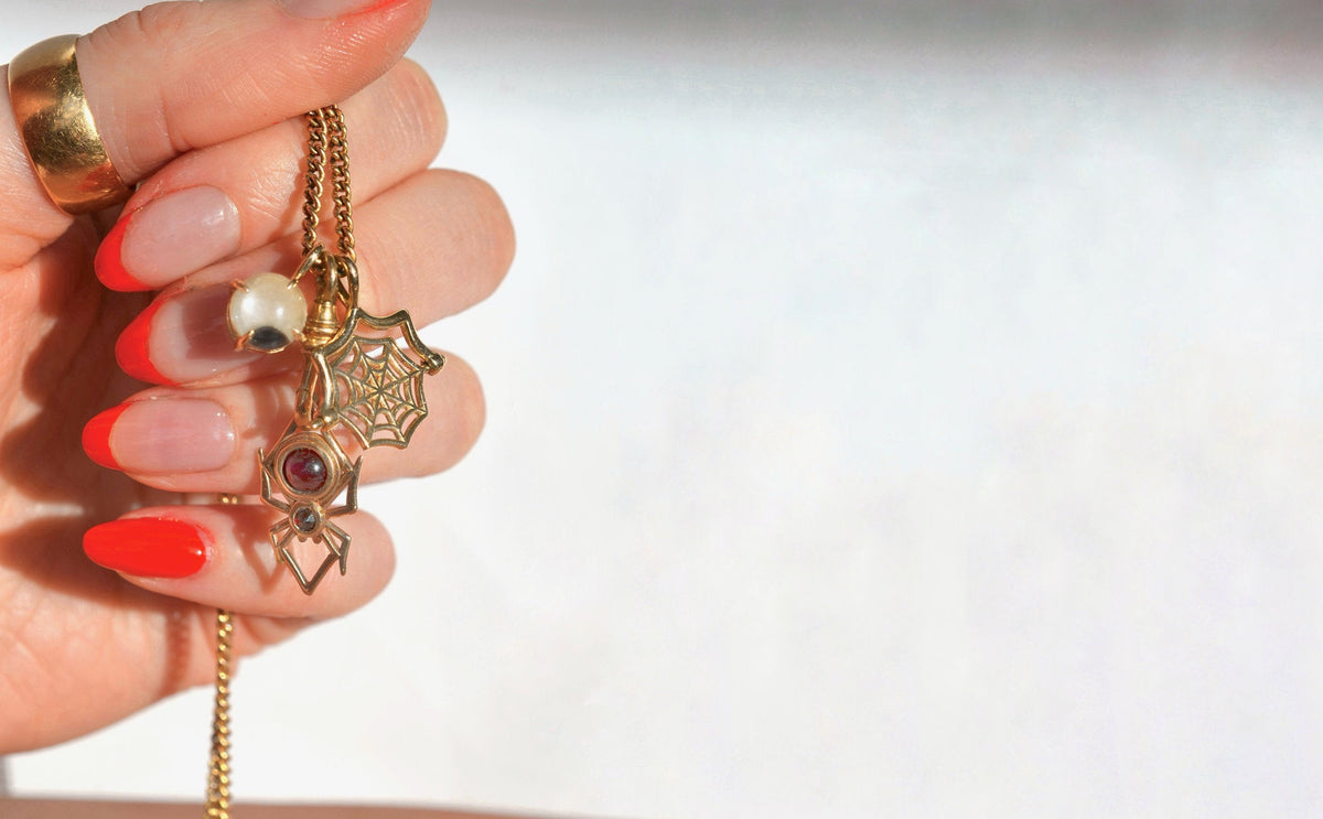 A hand with red and nude manicured nails holds a solid gold chain necklace featuring halloween-themed charms, including a spiderweb pendant, a spider silhouette charm, and a fine jewelry googly eye against a bright white background.