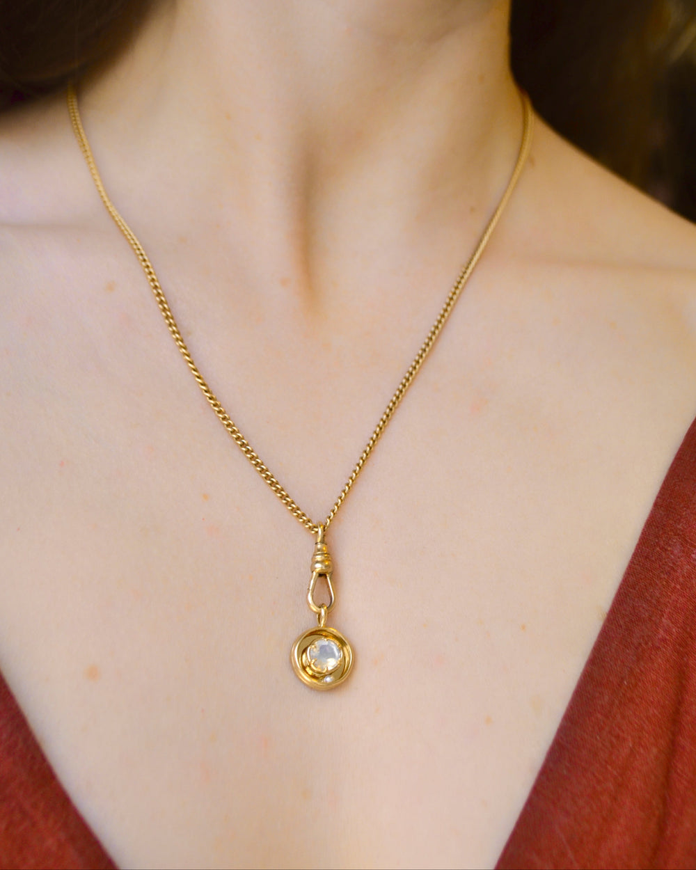 A close-up of a model wearing a kinetic rolling moonstone pendant on a delicate gold chain necklace, paired with a burnt-orange colored top.