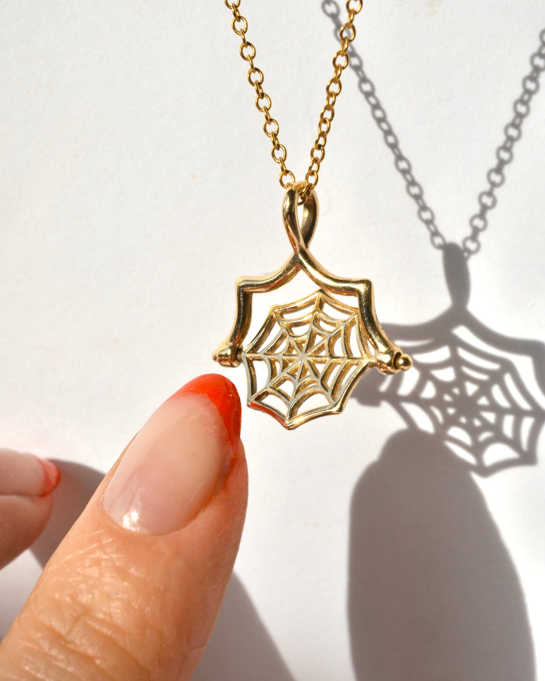 A gold spiderweb spinner pendant necklace being spun by a person's finger against a white background with a spiderweb shadow.